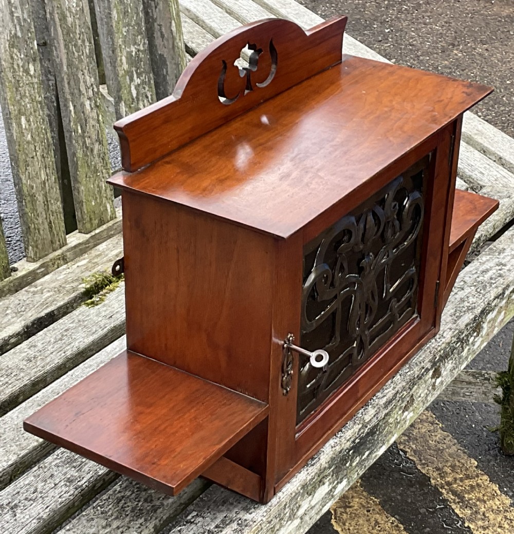 An Art Nouveau mahogany hanging wall cabinet featuring a central door with a decorative copper panel flanked by two side display shelves and topped with a pierced pediment. View VI