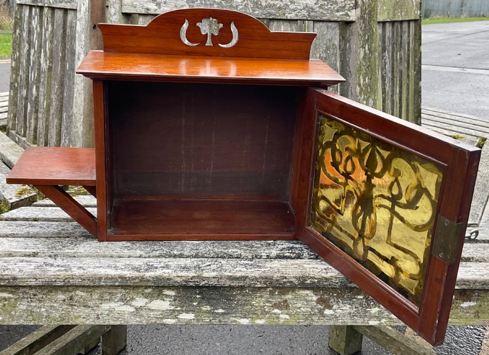 An Art Nouveau mahogany hanging wall cabinet featuring a central door with a decorative copper panel flanked by two side display shelves and topped with a pierced pediment. View VII