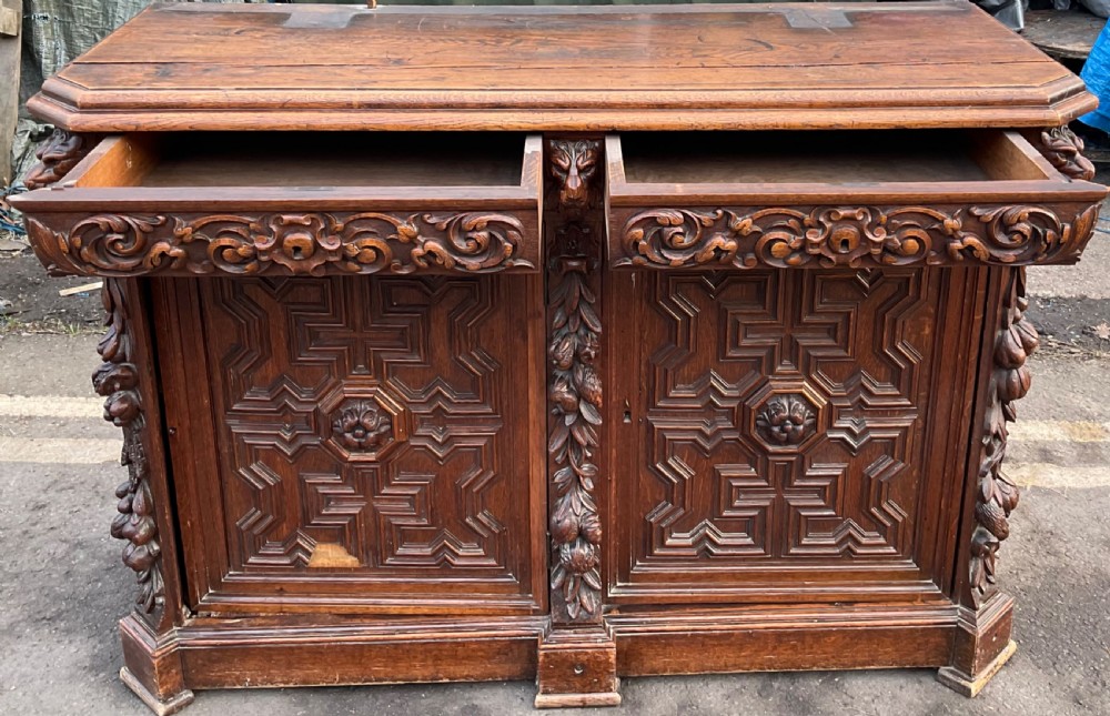 A dark Victorian carved oak sideboard featuring an ornate scrollwork cresting, tiered upper shelves, carved lion masks, and geometric panelled doors. View V