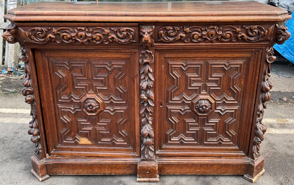 A dark Victorian carved oak sideboard featuring an ornate scrollwork cresting, tiered upper shelves, carved lion masks, and geometric panelled doors. View VI
