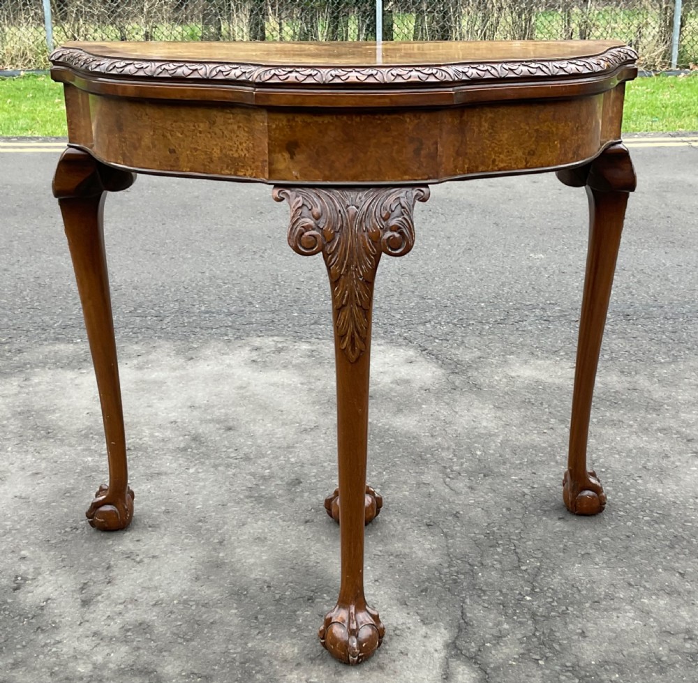 A semi-circular Queen Anne style burr walnut card table featuring a fold-over top, carved foliate border, and cabriole legs with ball-and-claw feet. View IV