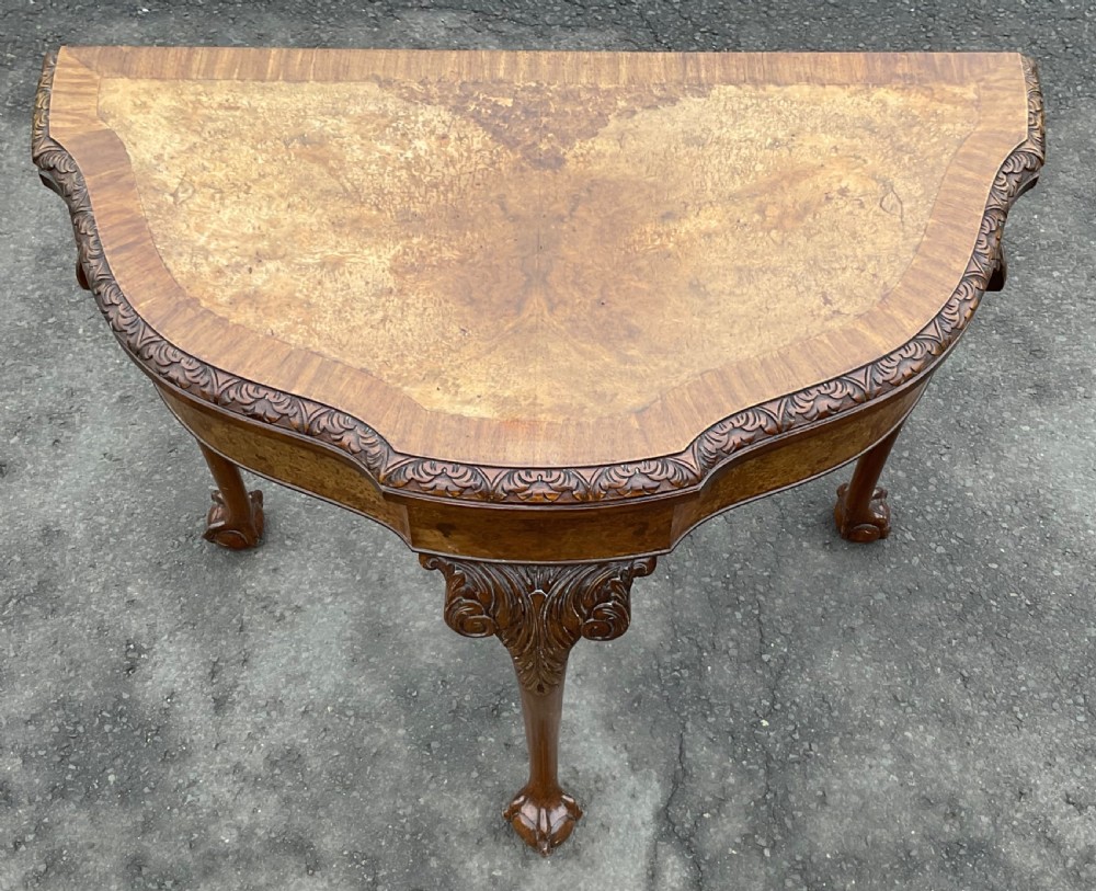 A semi-circular Queen Anne style burr walnut card table featuring a fold-over top, carved foliate border, and cabriole legs with ball-and-claw feet. View V