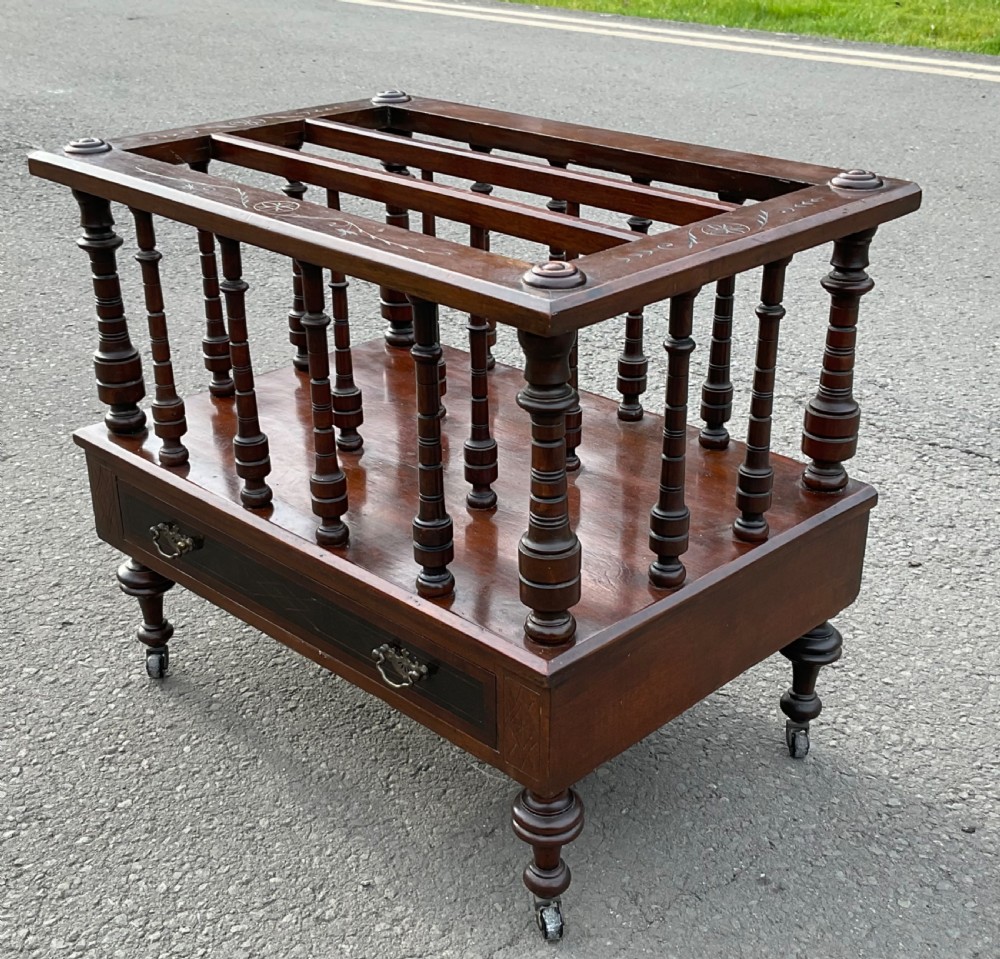 A Victorian dark mahogany Canterbury featuring vertical turned spindles, an inlaid top frame, a lower drawer with brass handles, and brass castors. View VI