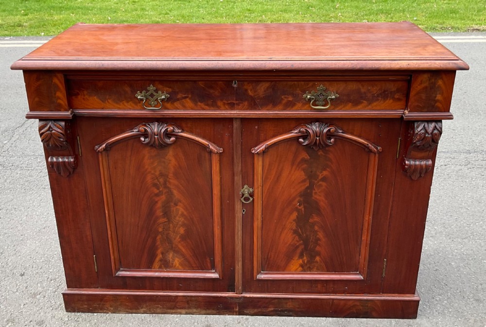 A Victorian mahogany two-door sideboard featuring flame mahogany door panels, carved scroll details, side corbels, and brass drawer handles.