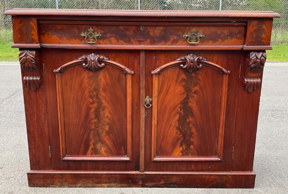 A Victorian mahogany two-door sideboard featuring flame mahogany door panels, carved scroll details, side corbels, and brass drawer handles. View II