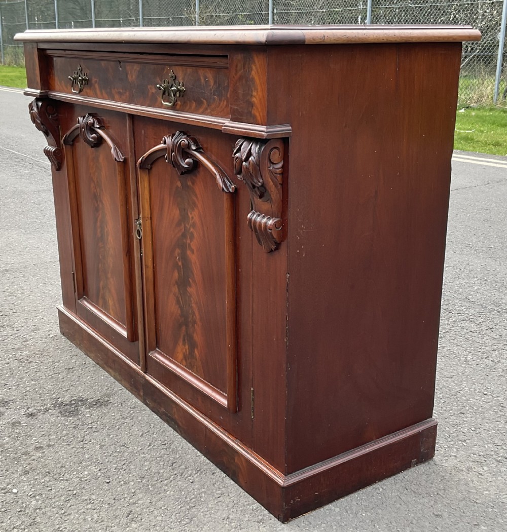 A Victorian mahogany two-door sideboard featuring flame mahogany door panels, carved scroll details, side corbels, and brass drawer handles. View III