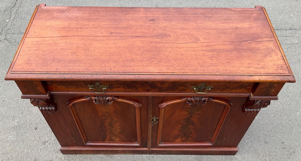 A Victorian mahogany two-door sideboard featuring flame mahogany door panels, carved scroll details, side corbels, and brass drawer handles. View IV