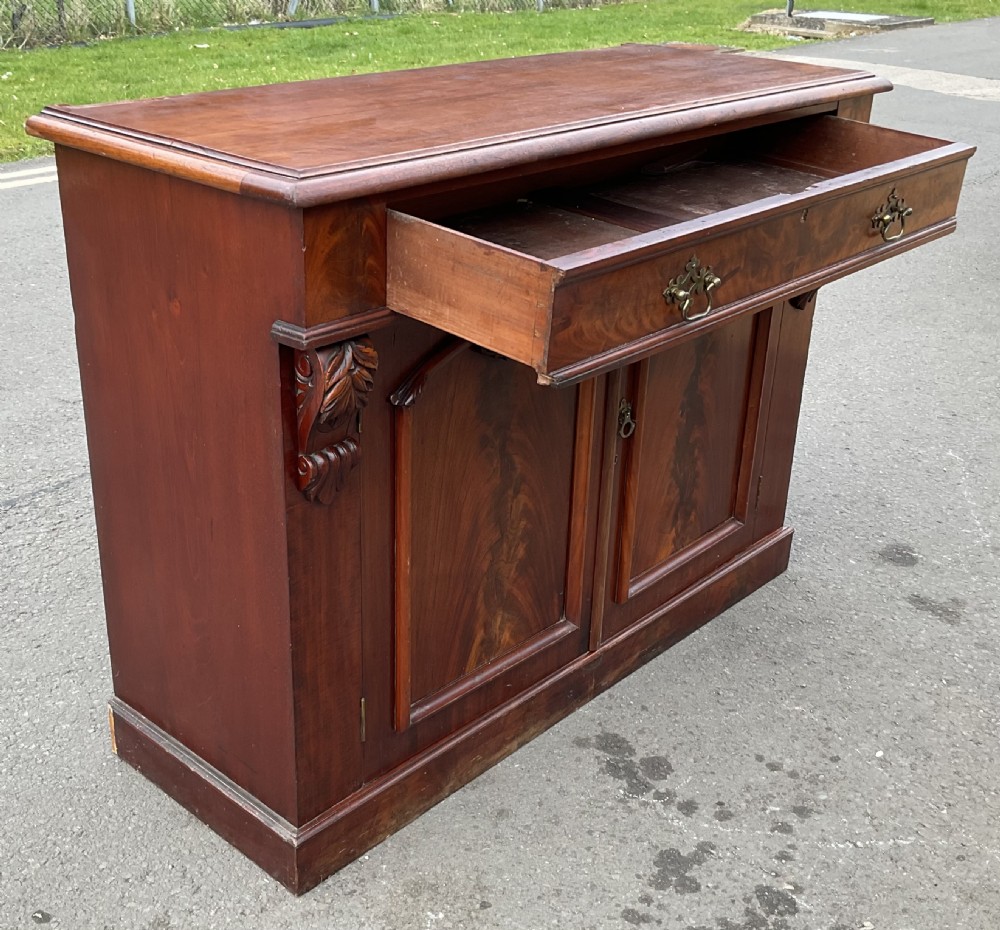 A Victorian mahogany two-door sideboard featuring flame mahogany door panels, carved scroll details, side corbels, and brass drawer handles. View VI