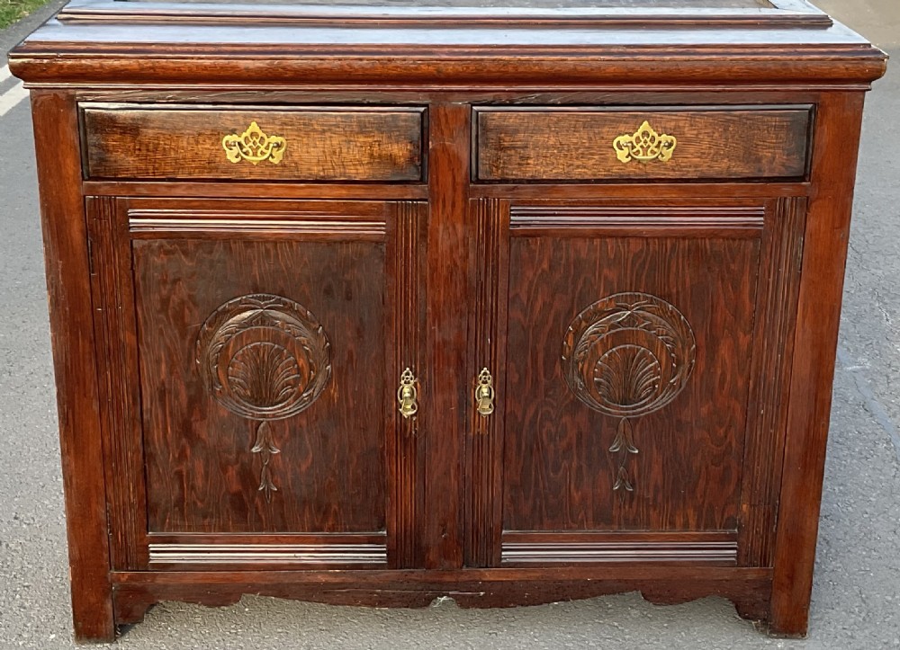 An Edwardian dark oak library bookcase featuring a carved top pediment, twin glazed upper doors, two drawers with brass handles, and carved lower cupboard doors. View VI