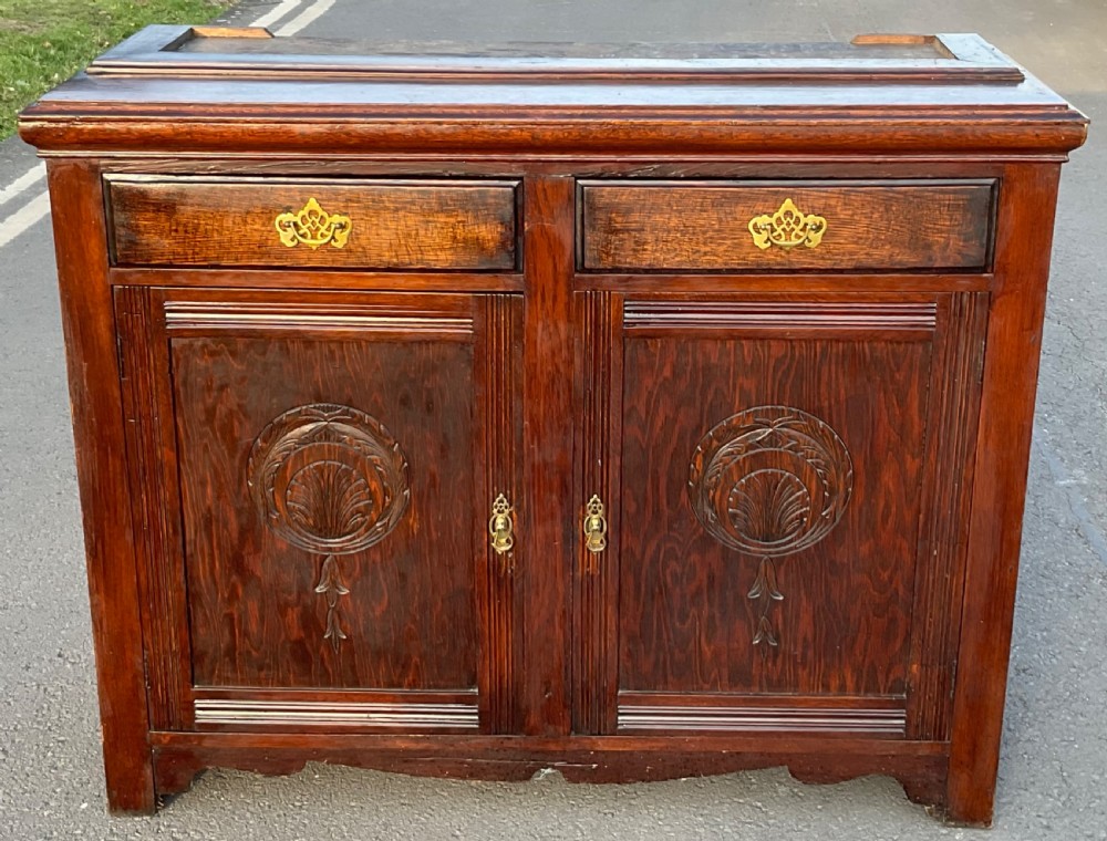 An Edwardian dark oak library bookcase featuring a carved top pediment, twin glazed upper doors, two drawers with brass handles, and carved lower cupboard doors. View VIII