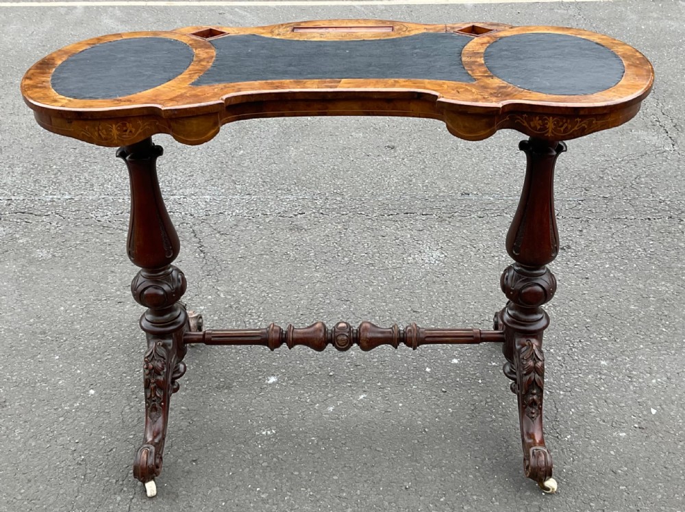 A Victorian burr walnut kidney-shaped writing table featuring an inset dark leather surface, floral marquetry inlay, carved turned supports, and a central stretcher base on castors.