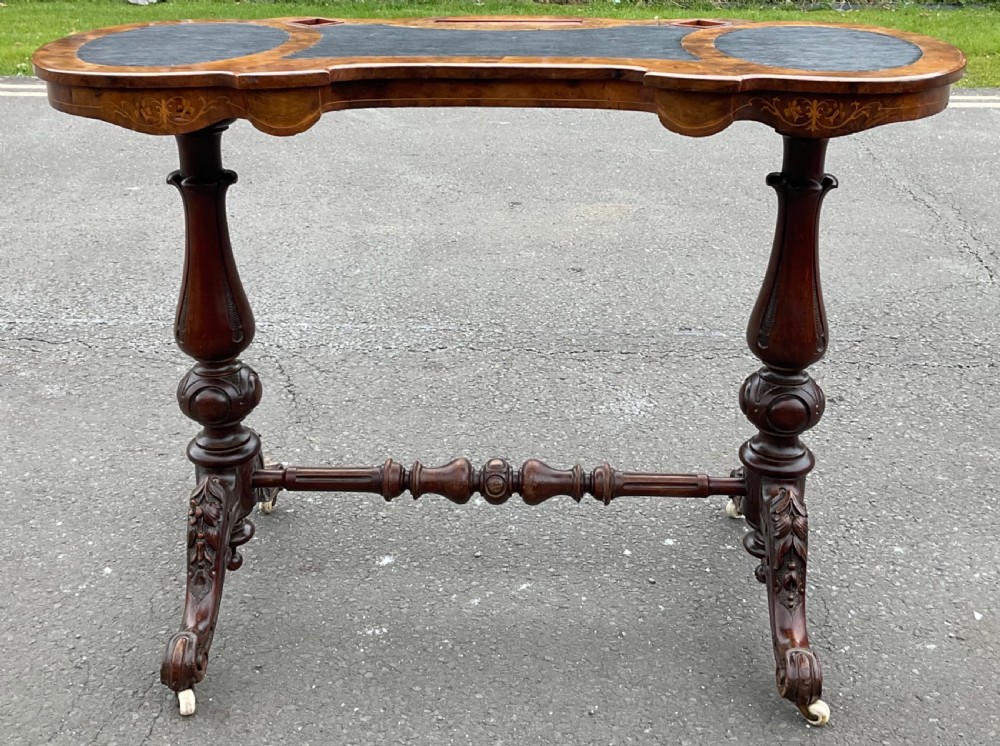 A Victorian burr walnut kidney-shaped writing table featuring an inset dark leather surface, floral marquetry inlay, carved turned supports, and a central stretcher base on castors. View II