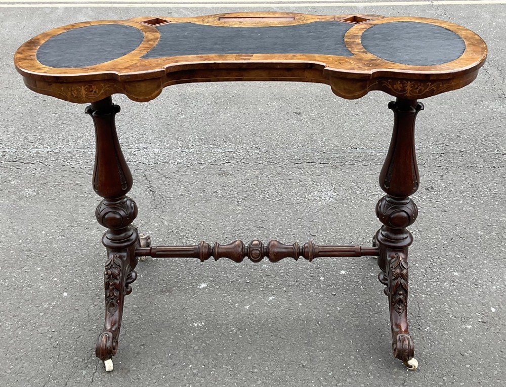 A Victorian burr walnut kidney-shaped writing table featuring an inset dark leather surface, floral marquetry inlay, carved turned supports, and a central stretcher base on castors. View III