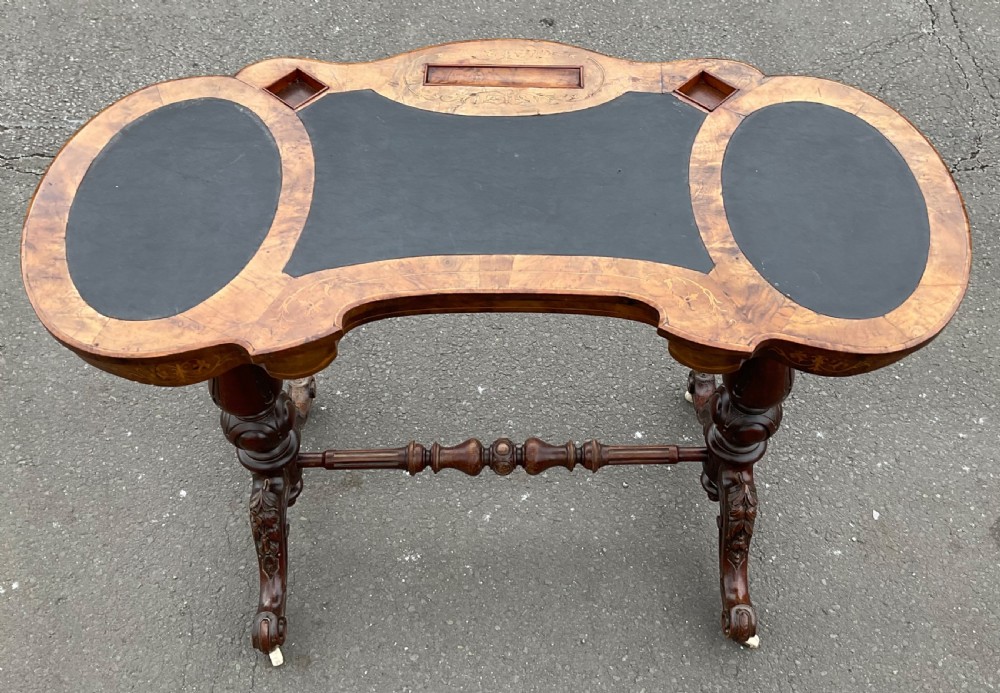 A Victorian burr walnut kidney-shaped writing table featuring an inset dark leather surface, floral marquetry inlay, carved turned supports, and a central stretcher base on castors. View IV