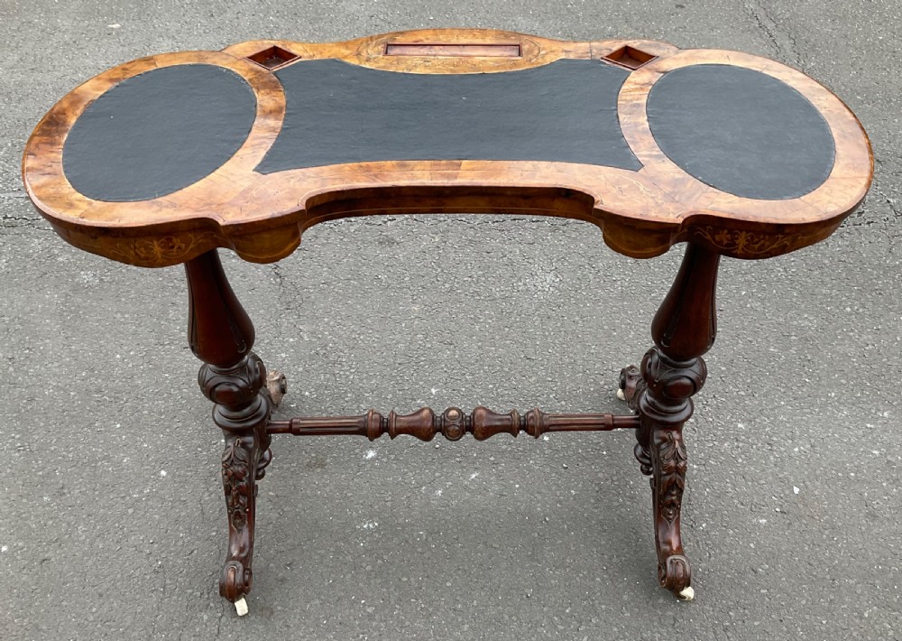 A Victorian burr walnut kidney-shaped writing table featuring an inset dark leather surface, floral marquetry inlay, carved turned supports, and a central stretcher base on castors. View V