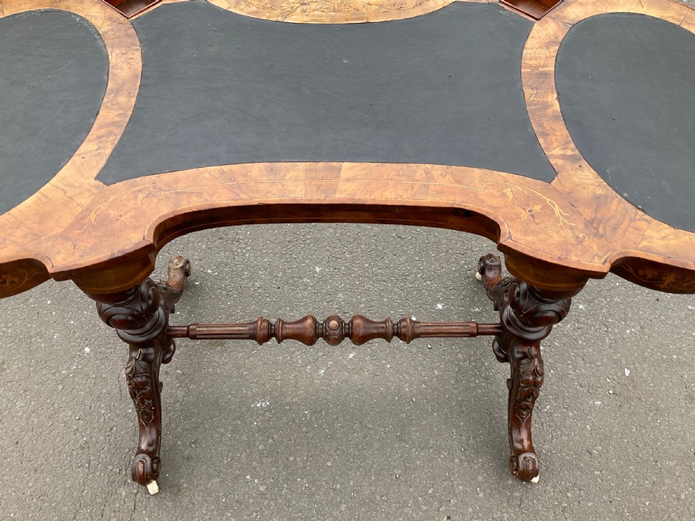A Victorian burr walnut kidney-shaped writing table featuring an inset dark leather surface, floral marquetry inlay, carved turned supports, and a central stretcher base on castors. View XIII