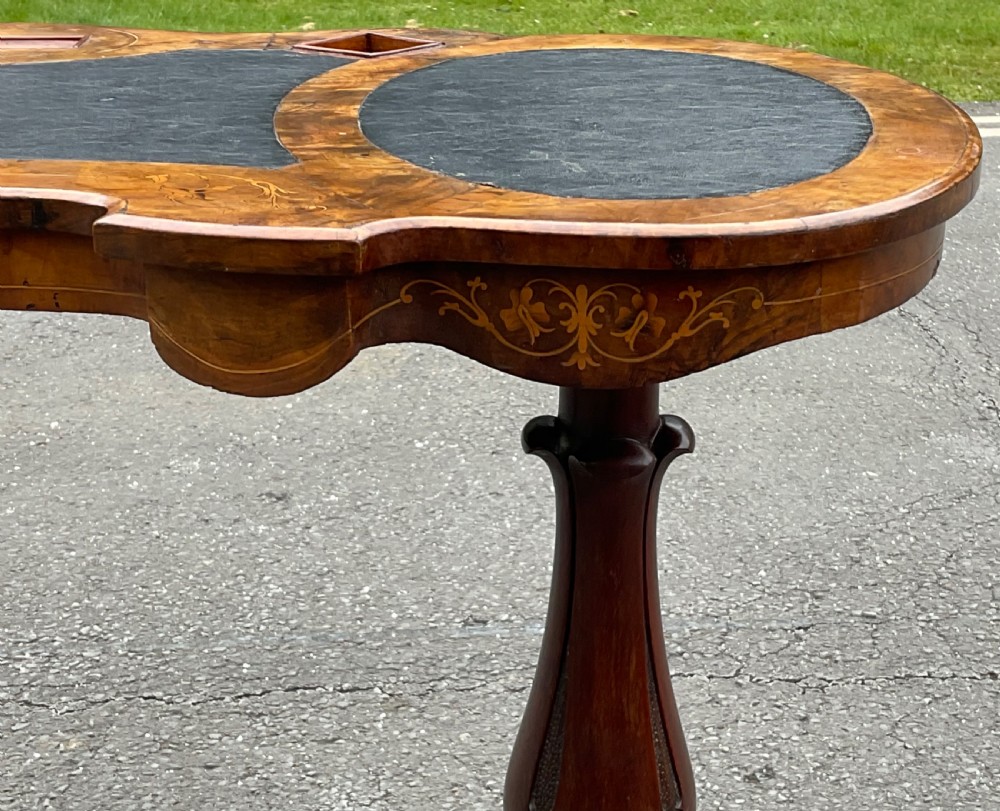A Victorian burr walnut kidney-shaped writing table featuring an inset dark leather surface, floral marquetry inlay, carved turned supports, and a central stretcher base on castors. View XIV