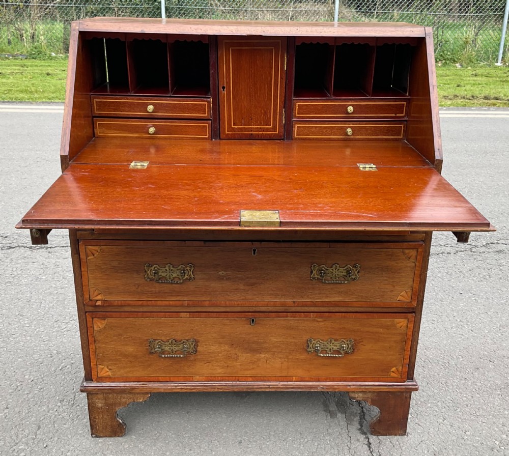 An Edwardian inlaid mahogany writing bureau featuring a sloped fall-front lid with a central oval marquetry motif above three drawers with brass handles. View VIII