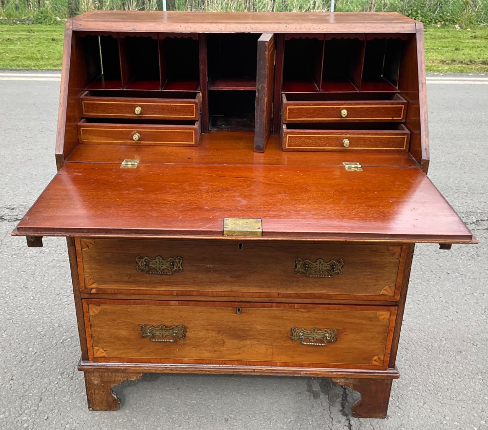 An Edwardian inlaid mahogany writing bureau featuring a sloped fall-front lid with a central oval marquetry motif above three drawers with brass handles. View IX
