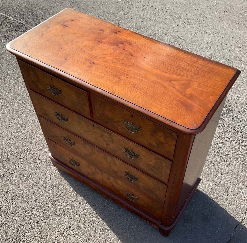 A Victorian burr walnut chest of drawers featuring a two-over-three drawer layout, decorative brass drop handles, and a solid plinth base. View VI