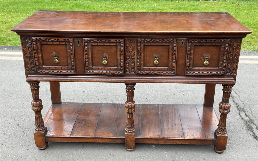 A dark carved oak Jacobean style dresser base featuring geometric panelled drawers, brass drop handles, central carved uprights, and a low potboard shelf supported by turned legs.