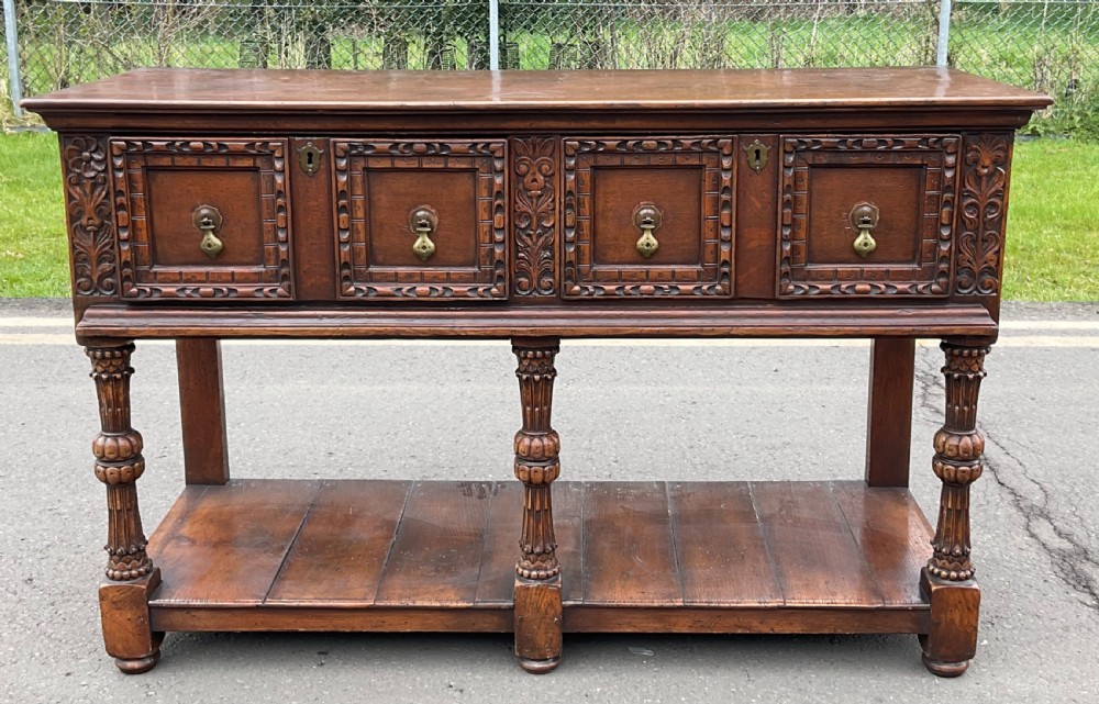 A dark carved oak Jacobean style dresser base featuring geometric panelled drawers, brass drop handles, central carved uprights, and a low potboard shelf supported by turned legs. View II