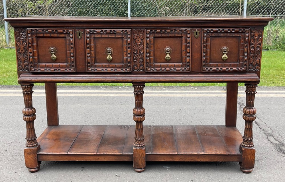 A dark carved oak Jacobean style dresser base featuring geometric panelled drawers, brass drop handles, central carved uprights, and a low potboard shelf supported by turned legs. View III