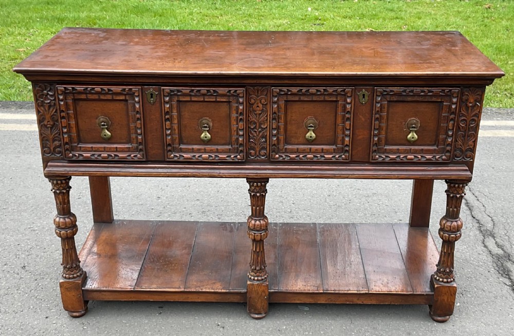 A dark carved oak Jacobean style dresser base featuring geometric panelled drawers, brass drop handles, central carved uprights, and a low potboard shelf supported by turned legs. View IV