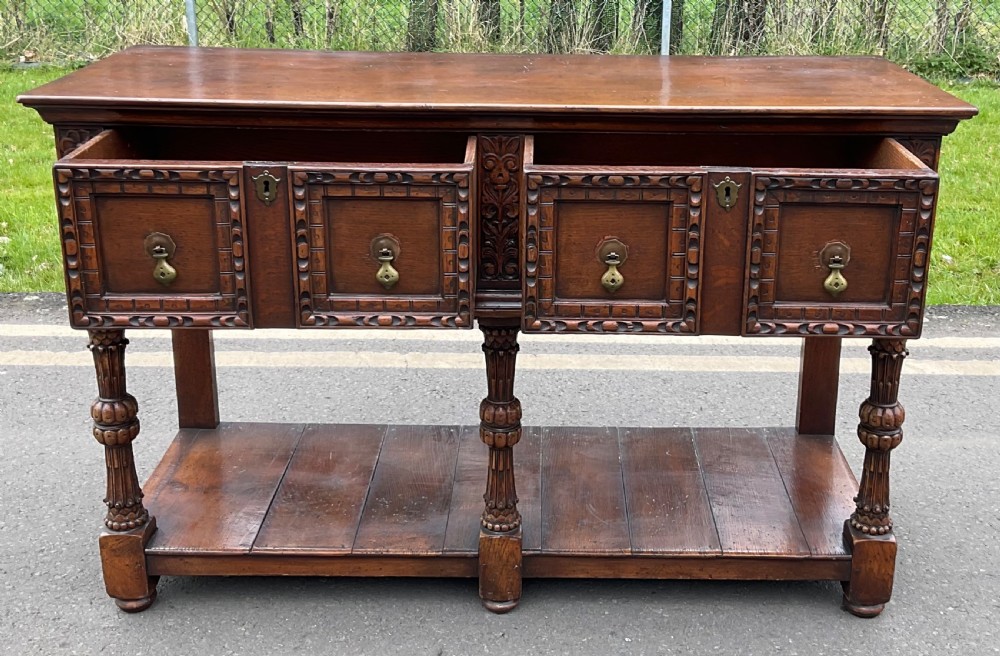 A dark carved oak Jacobean style dresser base featuring geometric panelled drawers, brass drop handles, central carved uprights, and a low potboard shelf supported by turned legs. View V