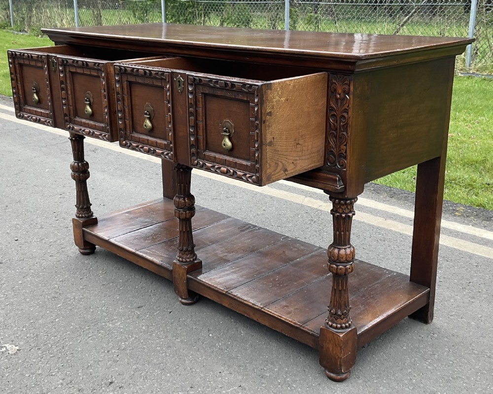 A dark carved oak Jacobean style dresser base featuring geometric panelled drawers, brass drop handles, central carved uprights, and a low potboard shelf supported by turned legs. View VIII
