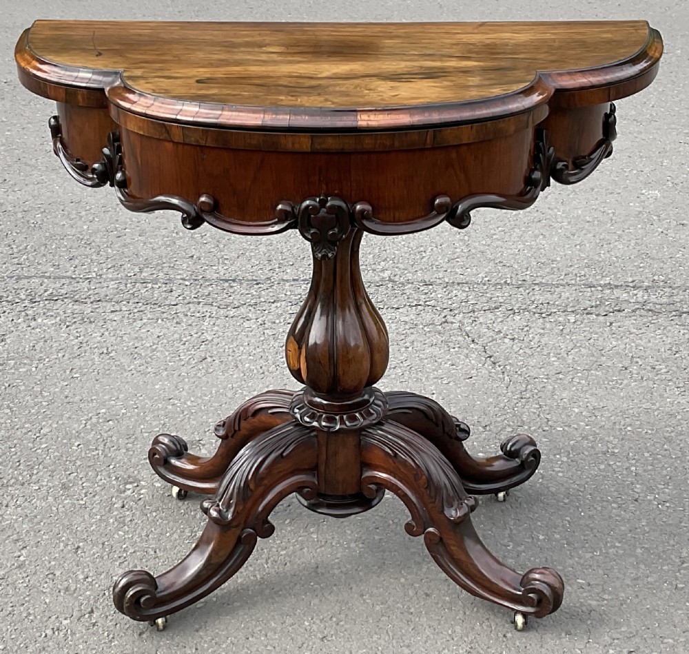 A Victorian rosewood fold-over card table featuring a shaped carved apron, bulbous turned pedestal, and four scrolled legs with brass castors.