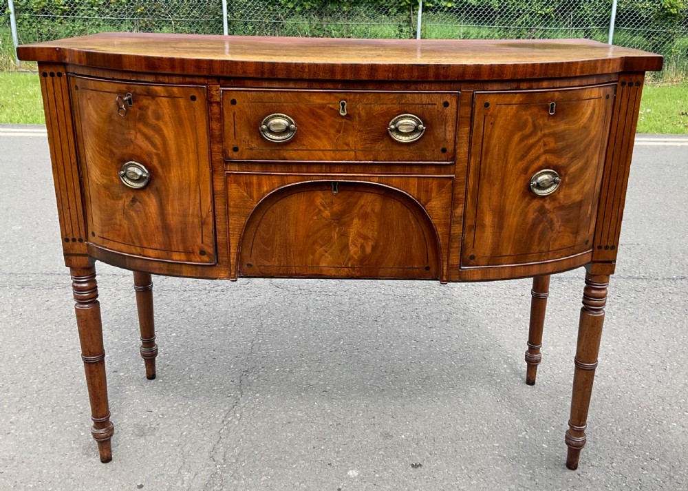 A Georgian bowfront mahogany sideboard featuring turned legs, brass oval drop handles, stringing inlay, and fluted corner pilasters.