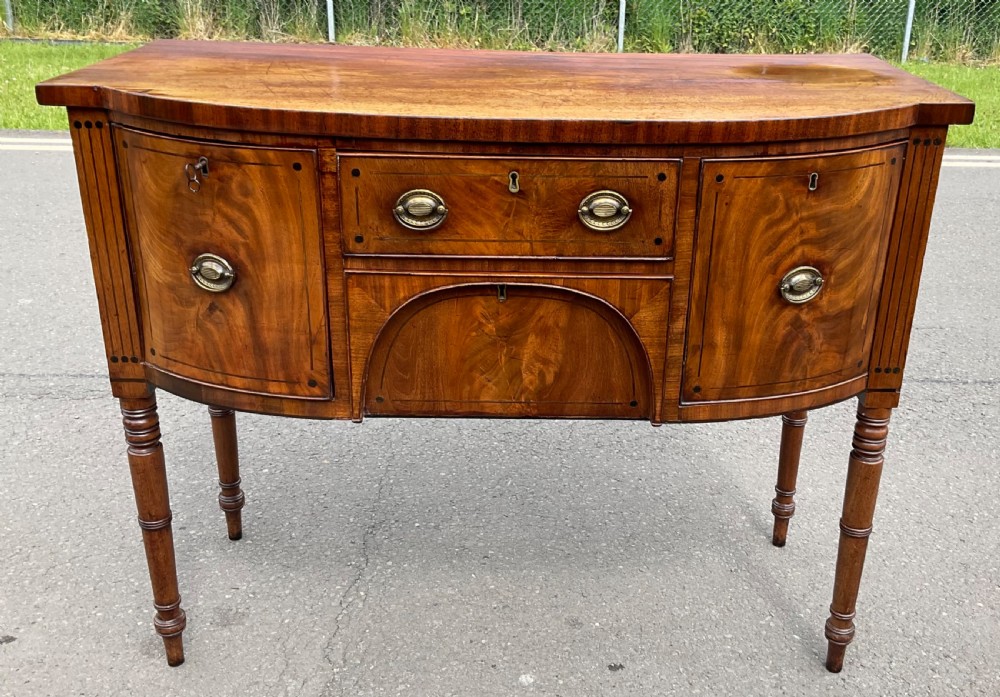 A Georgian bowfront mahogany sideboard featuring turned legs, brass oval drop handles, stringing inlay, and fluted corner pilasters. View II