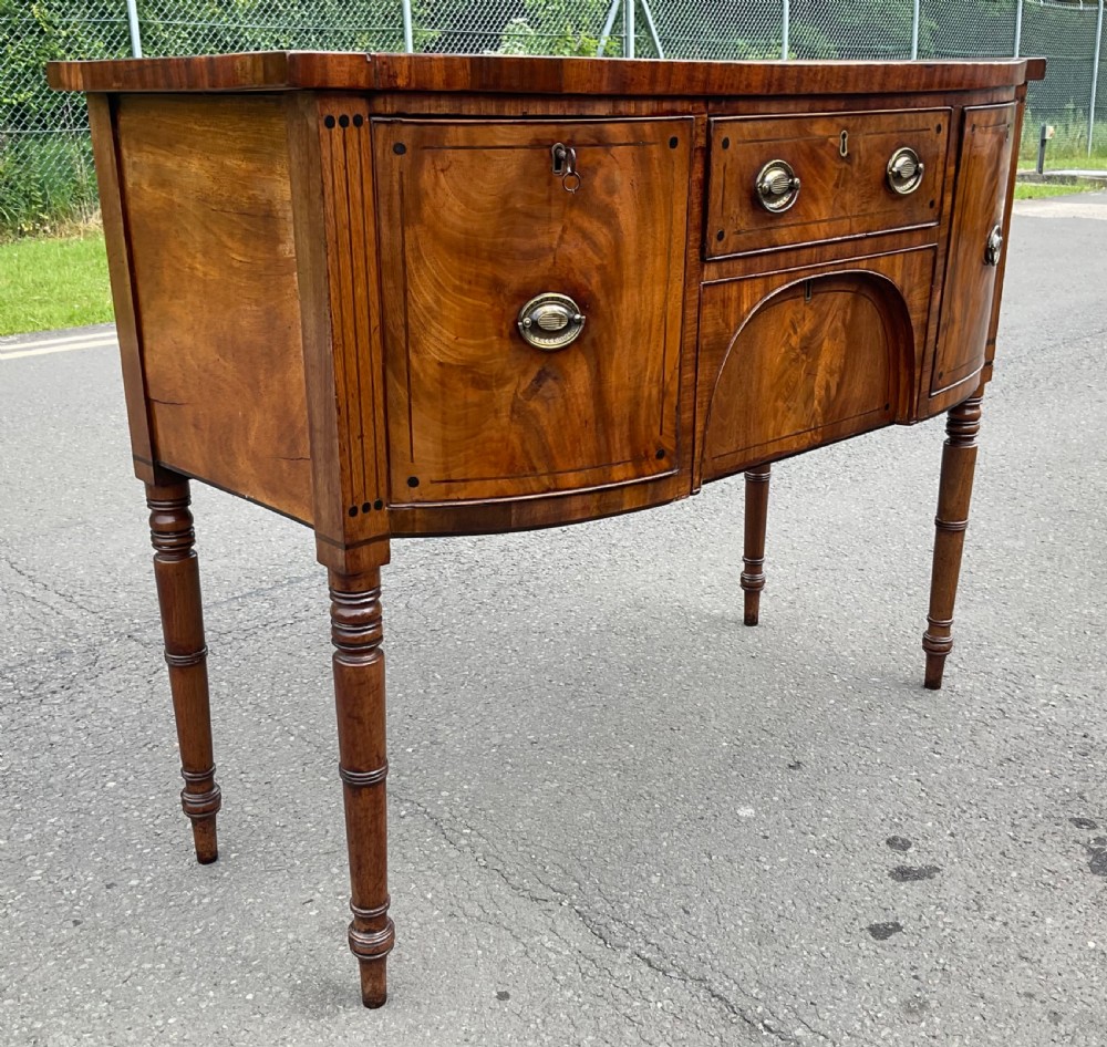A Georgian bowfront mahogany sideboard featuring turned legs, brass oval drop handles, stringing inlay, and fluted corner pilasters. View III