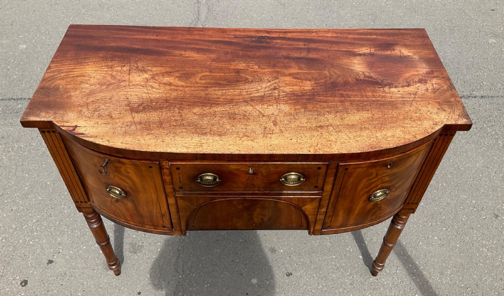 A Georgian bowfront mahogany sideboard featuring turned legs, brass oval drop handles, stringing inlay, and fluted corner pilasters. View V