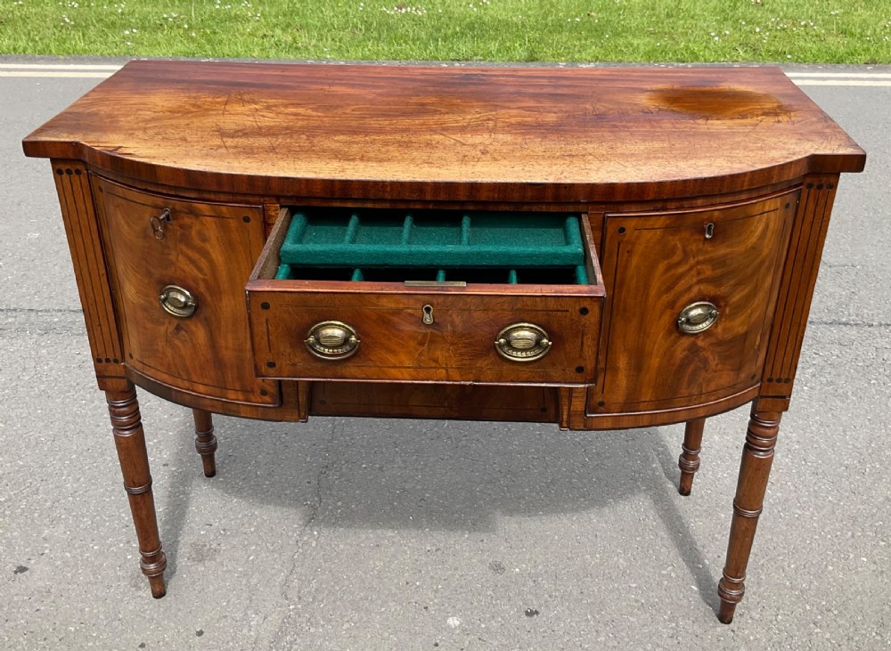 A Georgian bowfront mahogany sideboard featuring turned legs, brass oval drop handles, stringing inlay, and fluted corner pilasters. View VII