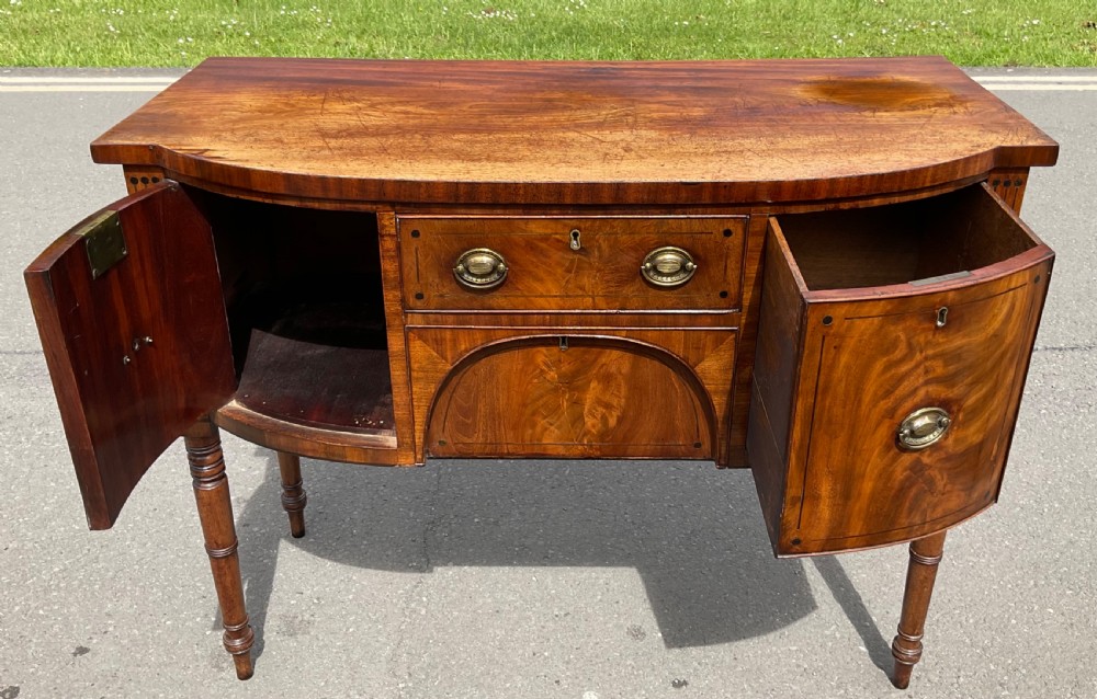 A Georgian bowfront mahogany sideboard featuring turned legs, brass oval drop handles, stringing inlay, and fluted corner pilasters. View VIII