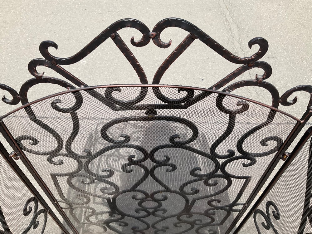 A tall black wrought iron folding fireguard with four hinged panels, decorative scrollwork, and fine wire mesh backing. View VI