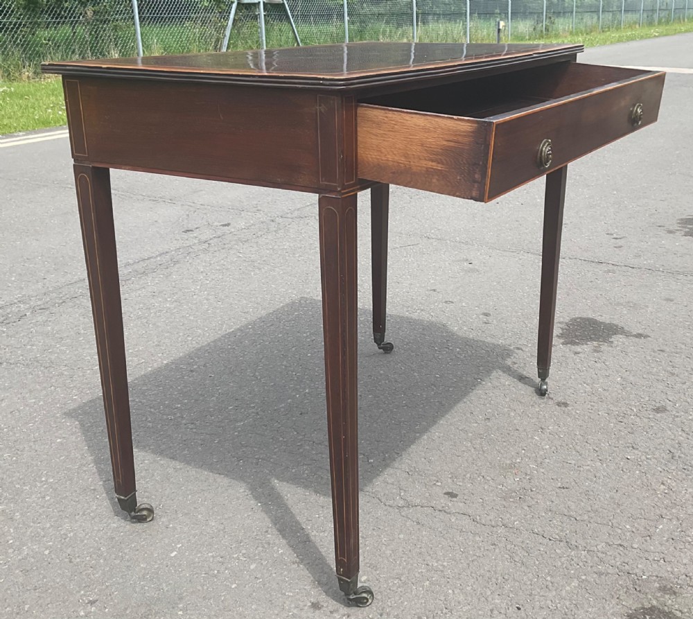 A Georgian mahogany writing table featuring boxwood string inlay on the rectangular top, a single drawer with twin brass ring handles, and square tapered legs on brass castors. View IX