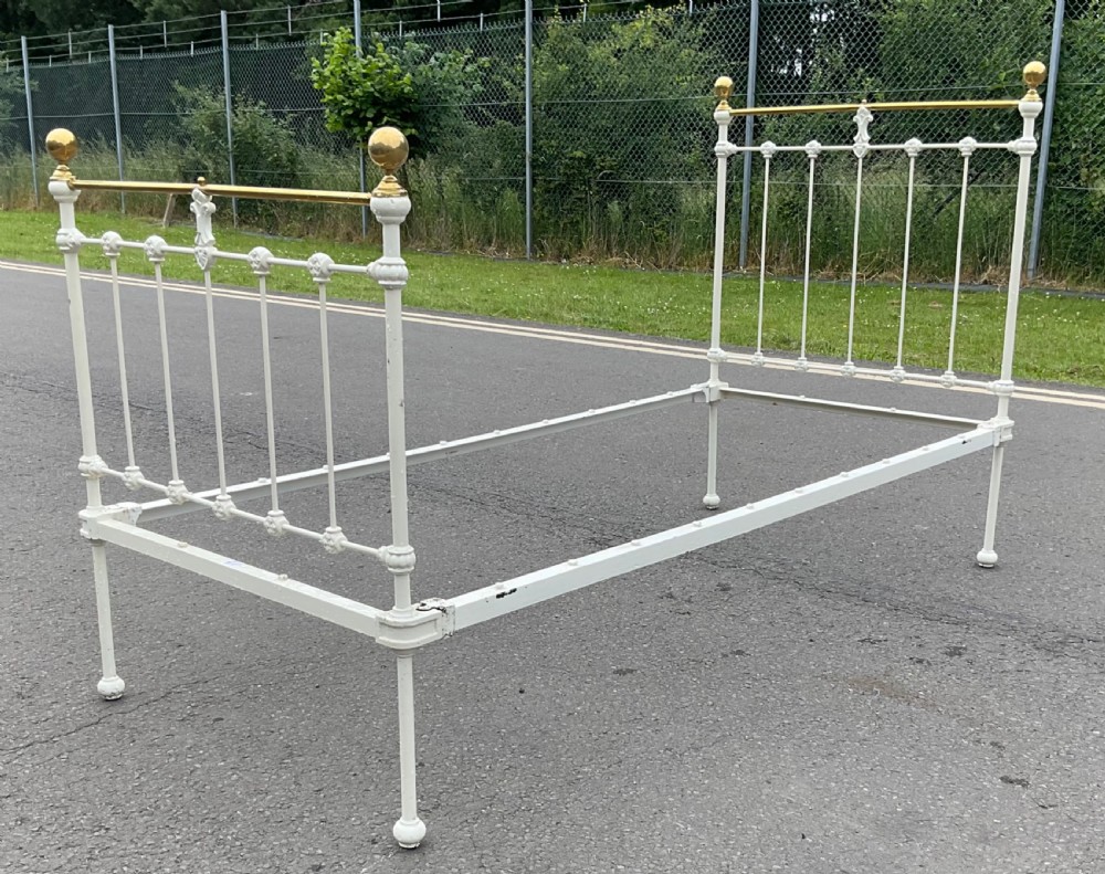 An off-white painted cast iron single bed frame featuring vertical spindles, decorative cast rosettes, central fleur-de-lis motifs, polished brass top rails, and brass spherical finials. View IV