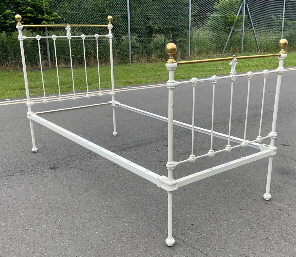 An off-white painted cast iron single bed frame featuring vertical spindles, decorative cast rosettes, central fleur-de-lis motifs, polished brass top rails, and brass spherical finials. View V
