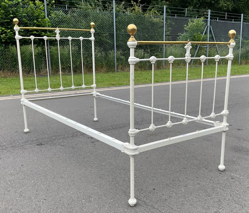 An off-white painted cast iron single bed frame featuring vertical spindles, decorative cast rosettes, central fleur-de-lis motifs, polished brass top rails, and brass spherical finials. View VI