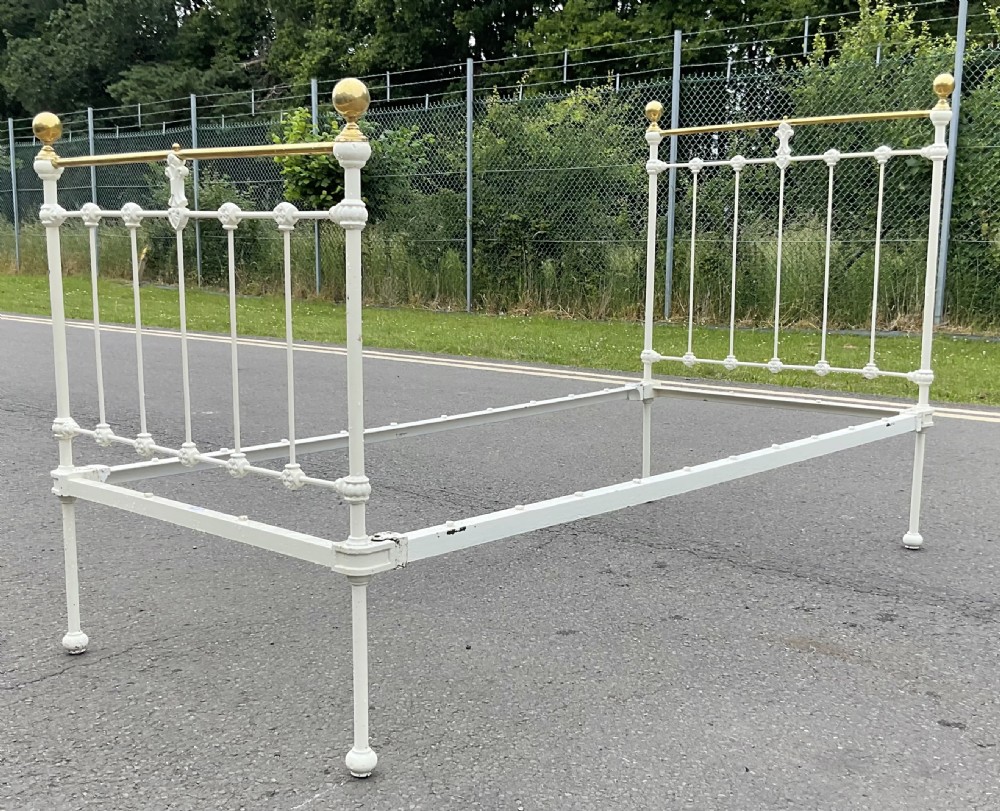 An off-white painted cast iron single bed frame featuring vertical spindles, decorative cast rosettes, central fleur-de-lis motifs, polished brass top rails, and brass spherical finials. View VII