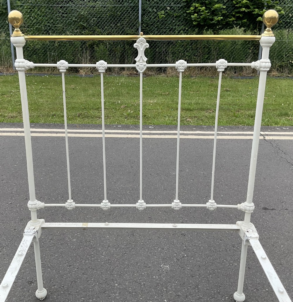 An off-white painted cast iron single bed frame featuring vertical spindles, decorative cast rosettes, central fleur-de-lis motifs, polished brass top rails, and brass spherical finials. View IX