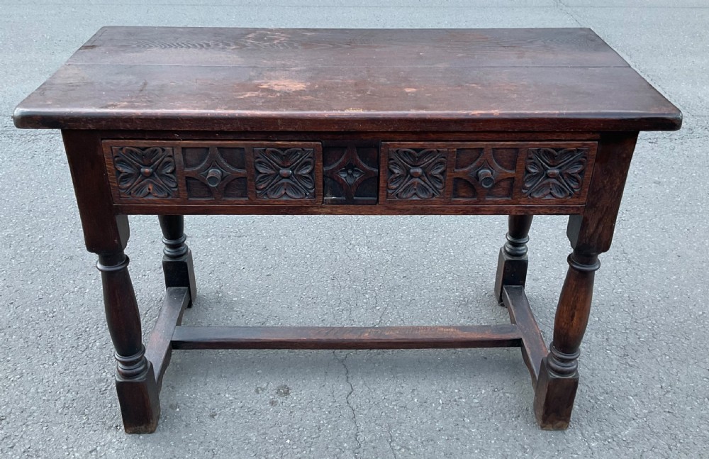A dark brown oak writing table featuring two carved panel drawers, turned front legs, and an H-stretcher base.