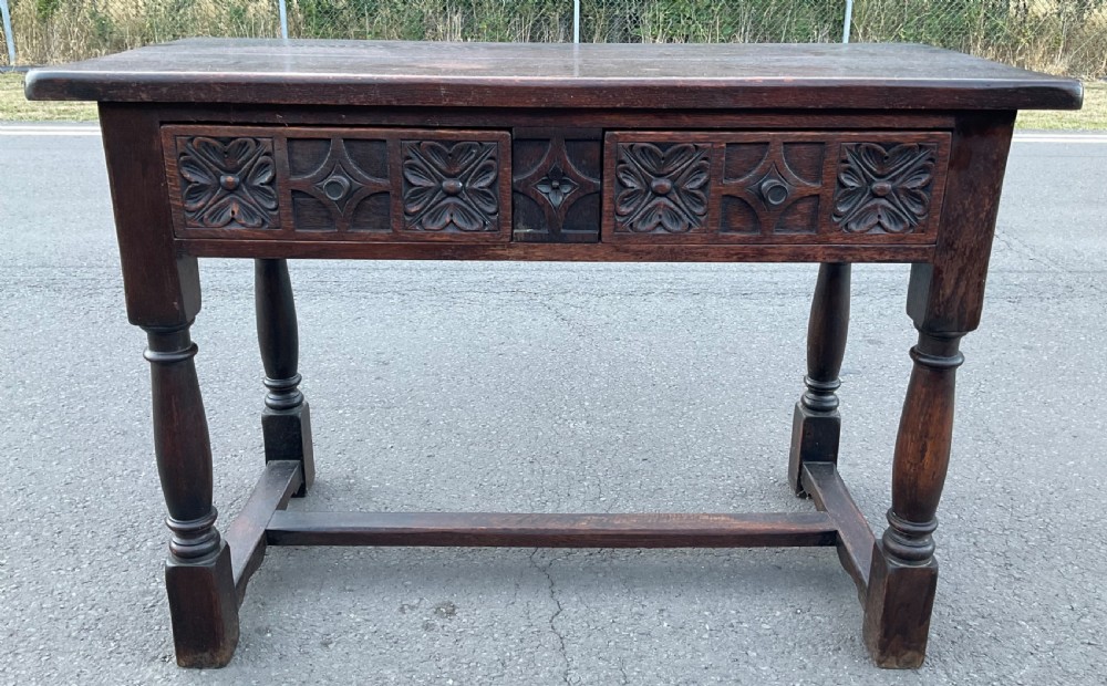 A dark brown oak writing table featuring two carved panel drawers, turned front legs, and an H-stretcher base. View II