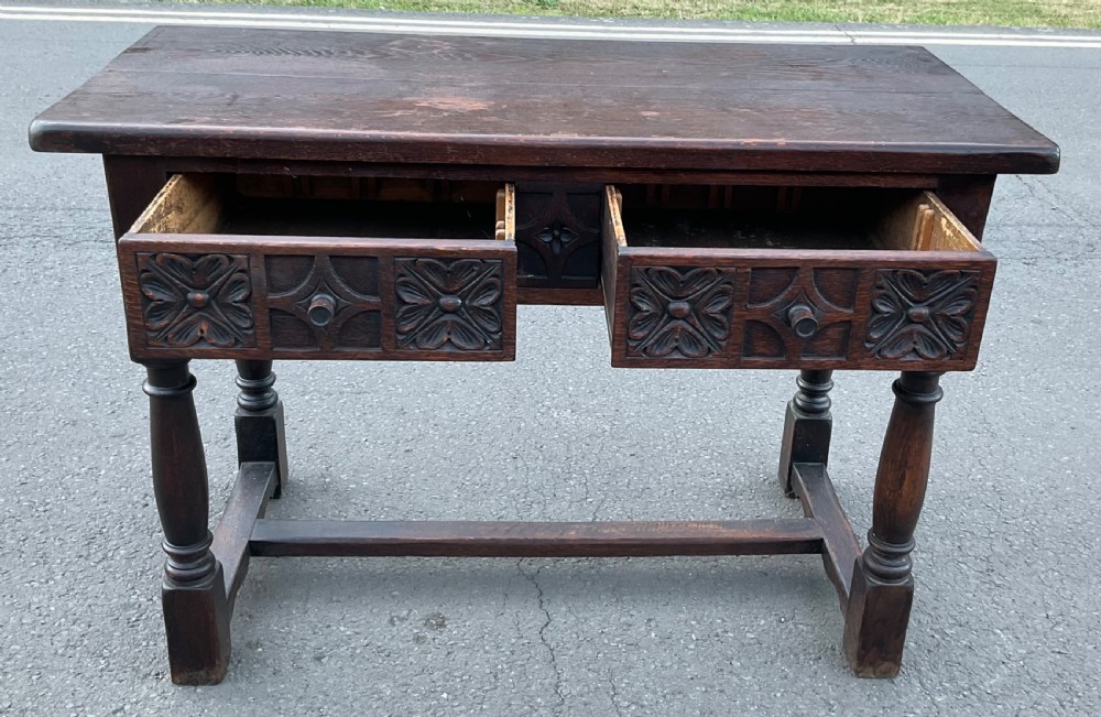 A dark brown oak writing table featuring two carved panel drawers, turned front legs, and an H-stretcher base. View III