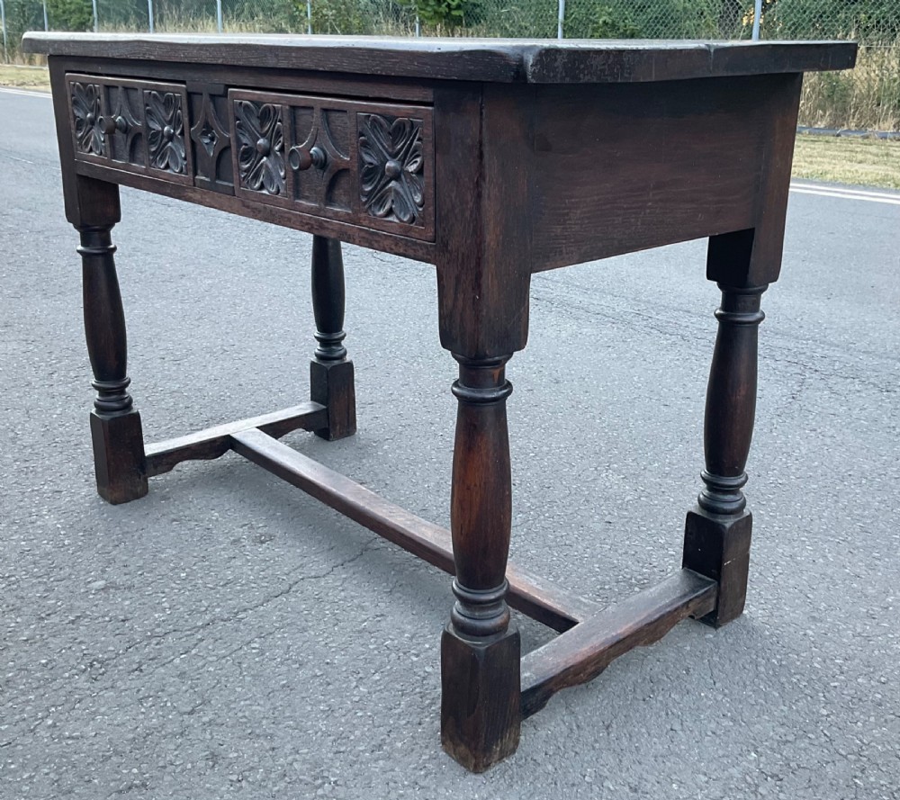A dark brown oak writing table featuring two carved panel drawers, turned front legs, and an H-stretcher base. View V