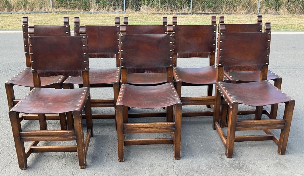 A set of eight dark oak dining chairs featuring thick brown leather slung seats and backrests trimmed with brass studs and leather fringe.
