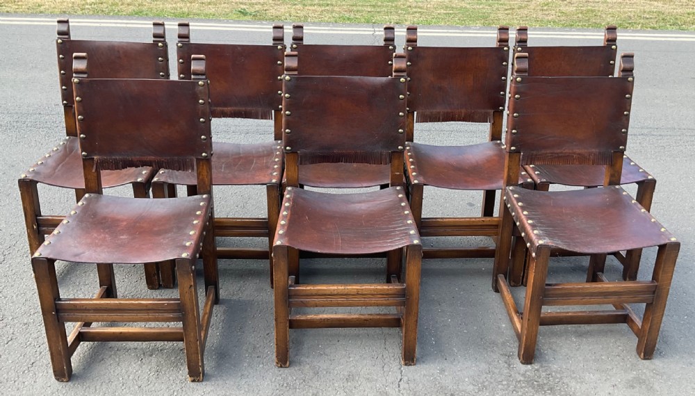 A set of eight dark oak dining chairs featuring thick brown leather slung seats and backrests trimmed with brass studs and leather fringe. View II